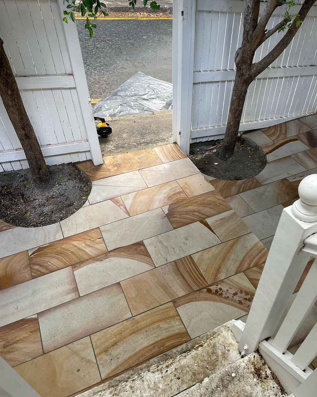 Sandstone-coloured pavers laid around two tree trunks beside a white gate
