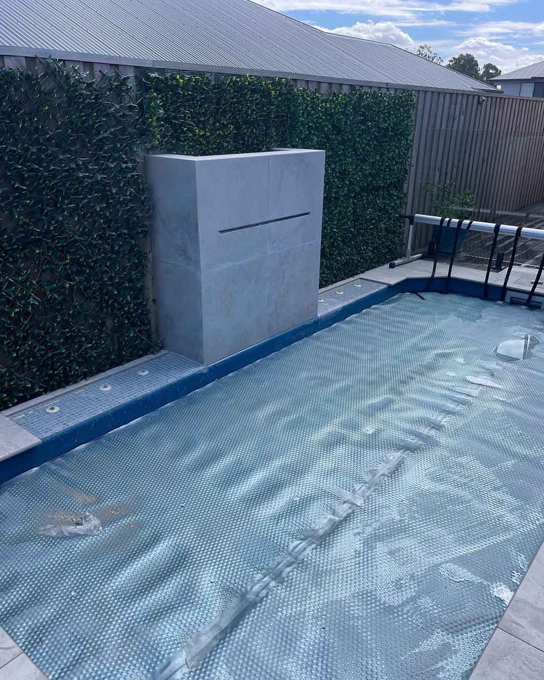 Covered pool with a grey tiled water feature wall in front of a hedge wall