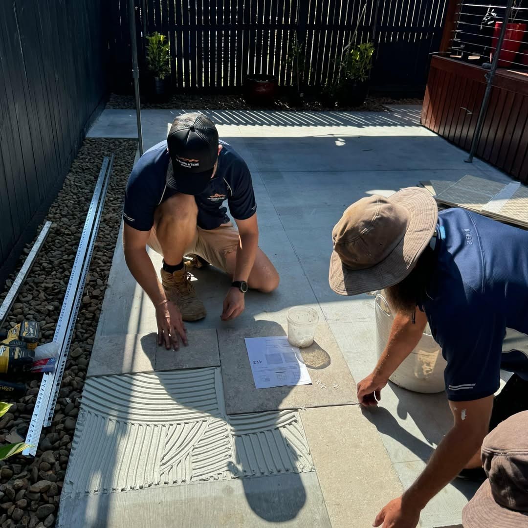 Two tradesmen laying large grey tiles on fresh adhesive outdoors