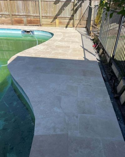 Pale stone pavers curving along a pool edge beside a garden fence