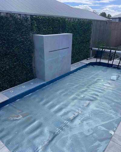 Covered pool with a tiled water feature wall and green hedge wall, wider view