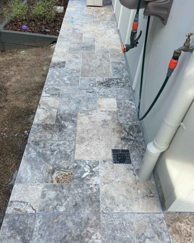 Grey stone pavers along the side of a house beside a downpipe and taps